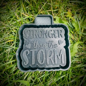 May include: A black plastic mould with the words "Stronger than the Storm" in a decorative font. The mould is sitting on a bed of green grass.