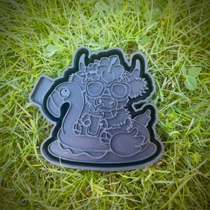 Può includere: Un tagliapasta in plastica nera a forma di mucca delle Highlands con occhiali da sole e una corona di fiori, che galleggia su un salvagente a forma di fenicottero e tiene una bevanda.