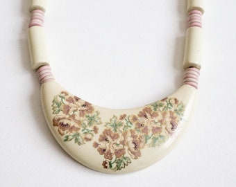 May include: A white ceramic necklace with a crescent-shaped pendant featuring a floral design in brown and green. The necklace is made of white and pink cylindrical beads.