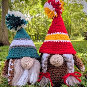 May include: Two crocheted gnome figures. The left gnome has a teal, white, and brown hat with a teal and white pom-pom and a long white beard. The right gnome has a red, yellow, and white hat with a multi-colored pom-pom and two braided pigtails.