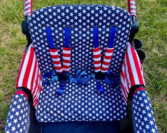 May include: A stroller seat cover and handle wrap set. The seat cover is navy blue with white stars, and the handle wrap is red, white, and blue. The seat straps have red and white striped covers. The stroller is on a grassy surface.
