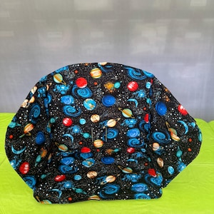 May include: A padded seat liner featuring a black background with a colorful space print. The fabric displays various planets, including Saturn with rings, and swirling blue galaxies scattered across a field of white stars and red celestial bodies.