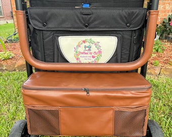May include: A black stroller with a brown leatherette seat and a brown leatherette cooler bag. The stroller has a black fabric canopy and a black fabric storage bag. The cooler bag has a zipper closure and a mesh pocket. The stroller has a footrest and two black wheels.