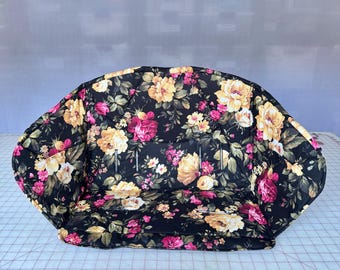 May include: A padded seat liner featuring a black background with a vibrant floral pattern of yellow, cream, and pink roses with green leaves. The liner includes integrated vertical slots for harness straps and a curved, contoured shape.