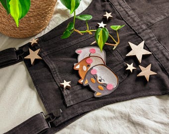 May include: Dark grey denim apron with adjustable straps and a large front pocket. Three embroidered patches of cartoon animals with pink cheeks are on the pocket. Wooden star-shaped decorations and a small plant are arranged on the apron.