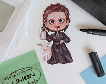 May include: A sticker depicting a cartoon woman in a dark dress, holding a bell and standing beside a lamb. The woman has blood on her mouth. The sticker is on a white surface with a pen, a small black object, and envelopes.