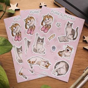 Op de afbeelding: Een set roze vinyl stickers met illustraties van katten in verschillende poses. De stickers maken deel uit van een vel met de titel "Pulla & Mörkö" en bevatten een kat die een boek leest, een kat met een vis en een kat met een pootprint.