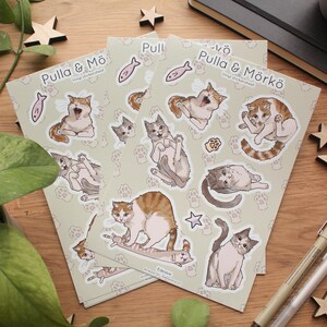 Op de afbeelding: Een set van drie vinyl sticker vellen met schattige cartoon katten in verschillende poses. De vellen zijn beige met poot afdruk ontwerpen en de tekst "Pulla & Mö" en "Pulla & Mörkö" in zwart. De stickers zijn perfect om een ​​toets van whimsy toe te voegen aan laptops, waterflessen, dagboeken en meer.