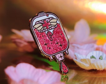 May include: A pink glitter enamel pin shaped like a blood bag with a red cross and wings. The pin has a gold outline and a dripping blood effect.