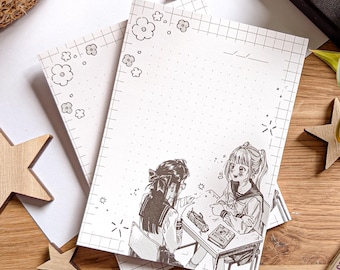 May include: White notepads with a grid pattern and floral accents. The notepads feature an illustration of two anime-style girls at a desk. Wooden star decorations are scattered around.