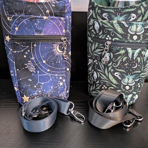 Puede incluir: Dos bolsas verticales de tela con correas ajustables. La bolsa de la izquierda presenta un estampado celestial azul oscuro con estrellas y lunas. La bolsa de la derecha tiene un fondo negro con patrones de polillas y helechos verdes.