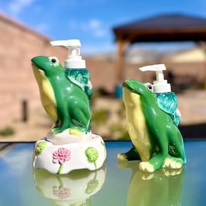May include: Two ceramic frog-shaped soap dispensers. The frogs are primarily green with yellow bellies and white pump tops. The base of one dispenser features pink flowers and green leaves. The dispensers are on a reflective surface.