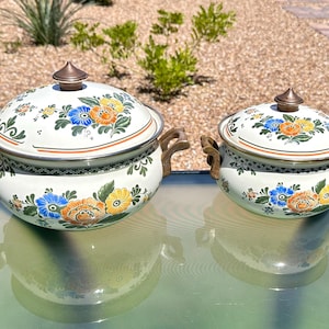 May include: Two white enamelware pots with lids, featuring floral designs in blue, orange, and yellow. The pots have bronze-colored handles and finials. The larger pot is approximately 10 inches in diameter, and the smaller pot is about 7 inches in diameter.