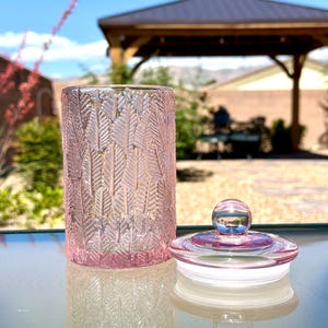 May include: A pink glass jar with a lid. The jar has a textured leaf pattern and is sitting on a glass table. The lid is clear with a small, round knob.