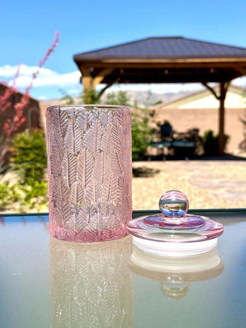 May include: A pink glass jar with a lid. The jar has a textured leaf pattern and is sitting on a glass table. The lid is clear with a small, round knob.