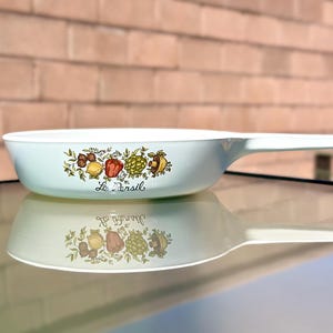 May include: A light blue vintage Le Persil casserole dish with a white handle. The dish features a colorful fruit and vegetable design. The dish is reflected on a glass surface.