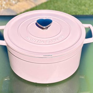 May include: A light pink Martha Stewart Dutch oven with a matching lid. The lid features a dark blue heart-shaped handle. The pot has two side handles and the brand name is embossed on the lid.