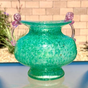 May include: A glass vase with a bulbous body featuring a speckled green and white pattern. The vase has a wide, flared rim and two translucent, light purple decorative handles attached to the sides, extending from the rim to the shoulder.