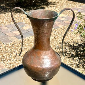 May include: A hammered copper vase featuring a wide, flared rim and two tall, slender curved handles extending from the neck to the shoulder. The metal surface displays a rich, weathered brown patina with subtle hints of green oxidation.