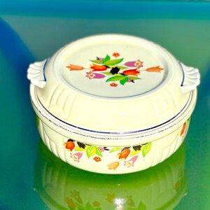 May include: A vintage, cream-colored casserole dish with a lid. The lid and the dish are decorated with a floral pattern in shades of orange, pink, and green. The dish has two handles and a silver trim.