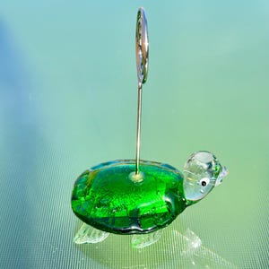 May include: A handcrafted glass turtle figurine with a vibrant green shell and clear head and flippers. A silver metal wire extends from the turtle's back, designed to hold a photo or note. The figurine is a decorative item.