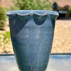 May include: A speckled blue ceramic vase with a wavy rim. The vase has a cylindrical shape and a textured surface. The blue color has variations, giving it a unique appearance. The vase is likely handmade and suitable for displaying flowers or as a decorative item.