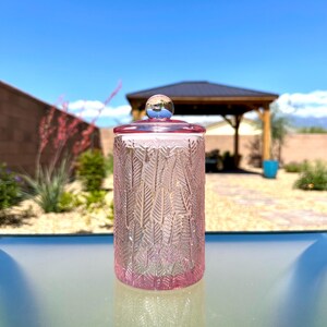 May include: A pink glass jar with a lid and a decorative leaf pattern. The jar is cylindrical and sits on a glass surface. The lid has a small, round, silver-colored knob. The background includes a gazebo and a clear blue sky.