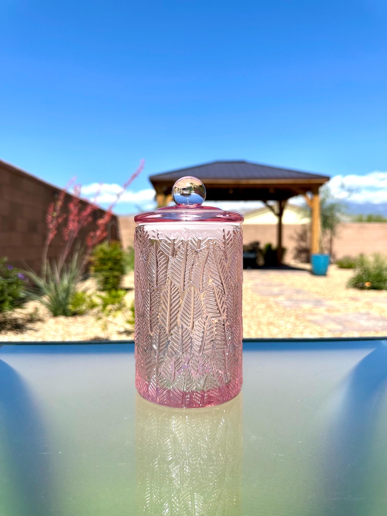 May include: A pink glass jar with a lid and a decorative leaf pattern. The jar is cylindrical and sits on a glass surface. The lid has a small, round, silver-colored knob. The background includes a gazebo and a clear blue sky.