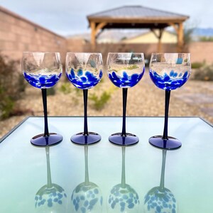 May include: Four clear wine glasses with blue speckled designs on the bowl and a dark blue stem. The glasses are arranged on a glass table, reflecting their image. The background features a gazebo and a residential yard.