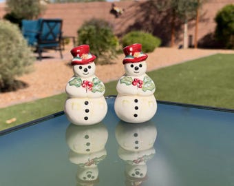 May include: Two ceramic snowman salt and pepper shakers featuring red top hats, green holly leaves with red berries on the chest, and three black buttons on the lower body. The figures have painted black eyes, noses, and mouths.