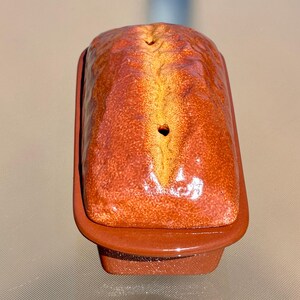 May include: A ceramic loaf pan with a lid. The pan and lid are a reddish-brown color with a textured surface. The lid has a heart-shaped hole. The pan is rectangular and appears to be handmade.