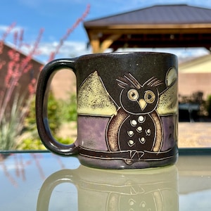 May include: A ceramic mug with a dark brown handle and body. The mug features a hand-painted owl design in shades of brown, purple, and yellow. The owl is centered on the mug, with a stylized landscape above and below it. The mug is sitting on a reflective surface.