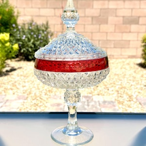 May include: A clear and red glass candy dish with a lid, featuring a diamond-cut pattern on the bowl and lid. The dish stands on a pedestal base and includes a pointed finial on top of the lid, showcasing a vibrant red band around the center.