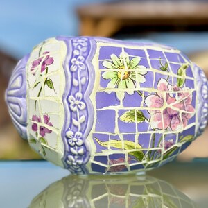 May include: A decorative, oval-shaped mosaic vase. The vase features a lavender background with a grid pattern, adorned with floral designs in pink, green, and white. The edges are decorated with white floral relief patterns. The vase is reflected on a glass surface.