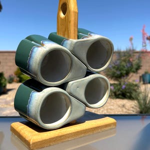May include: A set of four ceramic mugs in a teal and blue glaze, stacked on a wooden holder. The mugs have a unique shape and are arranged to showcase their design. The holder is made of light-colored wood, and the background is a clear blue sky.