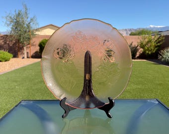May include: A round, translucent pink glass plate featuring an embossed floral pattern of roses and leaves. The plate has a scalloped edge and is displayed upright on a black plastic stand, showcasing its delicate texture and color.
