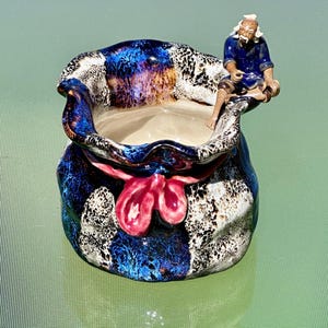 May include: A ceramic decorative bowl with a blue, black, and white pattern, tied with a pink bow. A small figurine of a man in blue clothing sits on the edge of the bowl. The bowl has a glossy finish and a light interior.