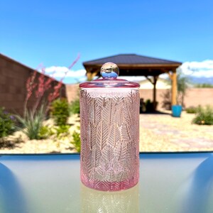 May include: A pink glass jar with a lid and a silver knob. The jar has a repeating leaf pattern. The lid and the base of the jar are a darker shade of pink. The background is a bright blue sky.