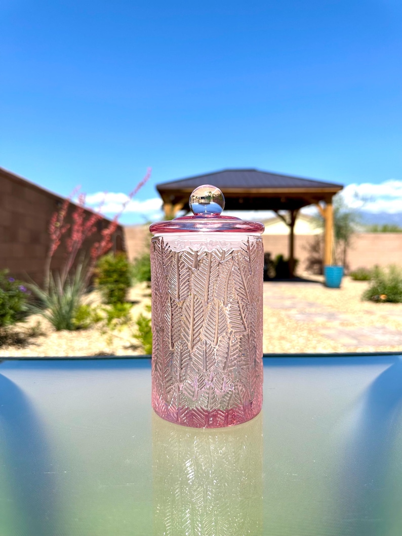 May include: A pink glass jar with a lid and a silver knob. The jar has a repeating leaf pattern. The lid and the base of the jar are a darker shade of pink. The background is a bright blue sky.