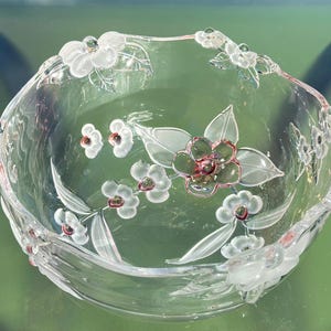 May include: Clear glass bowl with floral design. The bowl features etched white flowers with red and green accents. The rim of the bowl is also decorated with white flowers. The bowl is sitting on a green surface.