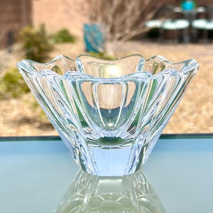 May include: A clear, faceted glass bowl with a scalloped rim. The bowl has a geometric design with vertical lines and a square base. The bowl is sitting on a reflective surface, creating a mirrored effect.