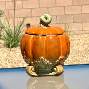 May include: A glossy orange ceramic pumpkin-shaped lidded jar with a light green stem handle. The base features decorative leaf-like carvings and scroll-shaped feet in a muted green tone, providing a stable foundation for the rounded vessel.