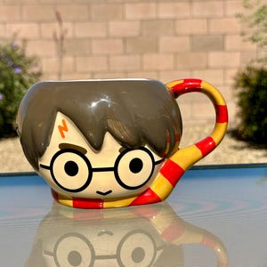 May include: A ceramic mug shaped like the head of a character with dark brown hair, round black glasses, and a lightning bolt scar on the forehead. The handle is designed as a scarf with alternating horizontal red and yellow stripes.