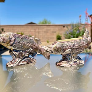 May include: Two ceramic fish figurines with a glossy, multi-colored glaze featuring shades of brown, green, and gold. Each fish is sculpted in a swimming position, mounted on a base that resembles splashing water, with one fish featuring an open top.
