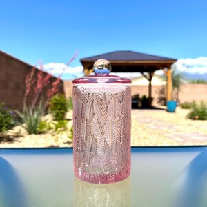 May include: A pink glass jar with a lid and a silver knob. The jar has a textured pattern of vertical leaves. The lid and the base of the jar are a darker shade of pink. The jar is sitting on a glass surface.