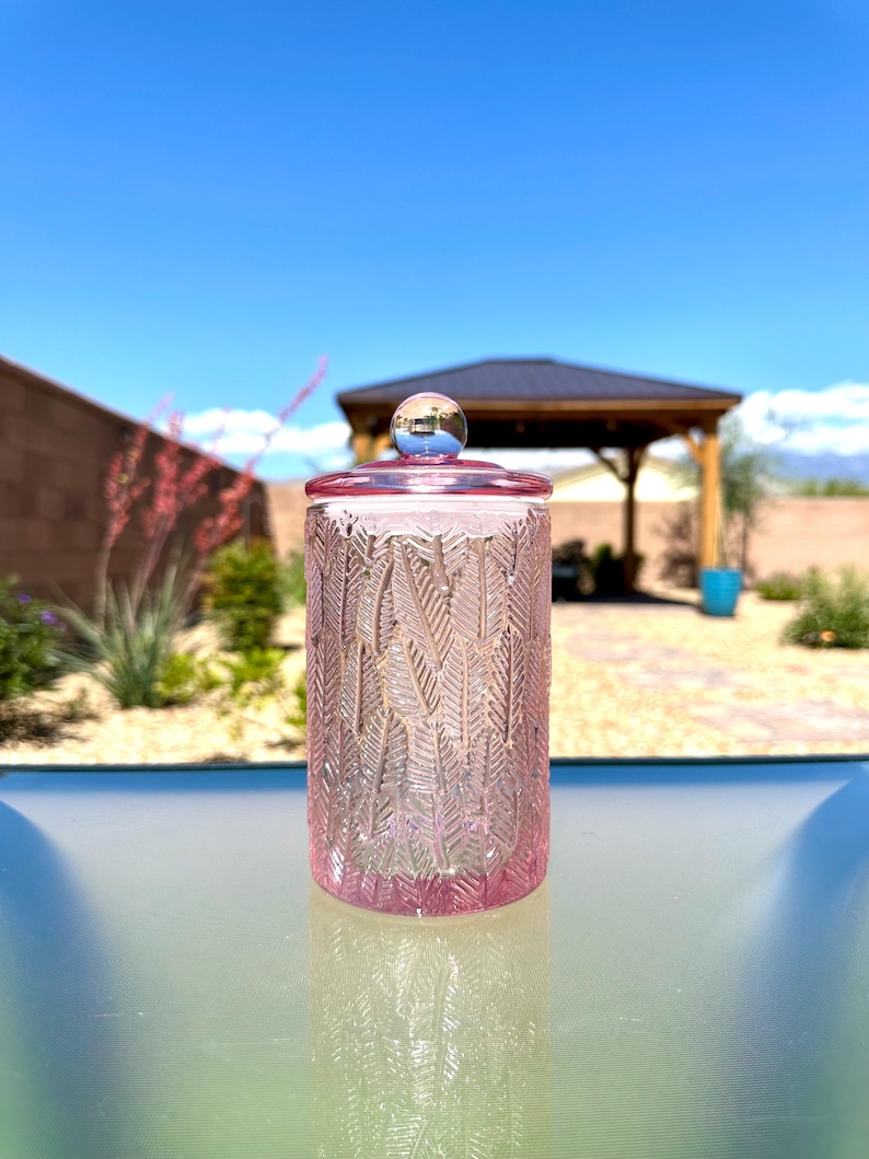 May include: A pink glass jar with a lid and a silver knob. The jar has a textured pattern of vertical leaves. The lid and the base of the jar are a darker shade of pink. The jar is sitting on a glass surface.