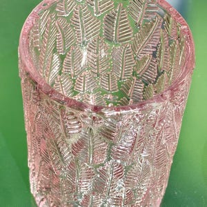 May include: A clear, pink glass tumbler with a detailed leaf pattern. The cylindrical glass has a slightly flared rim and is designed with a repeating embossed fern-like motif. The glass is suitable for beverages and decorative use.
