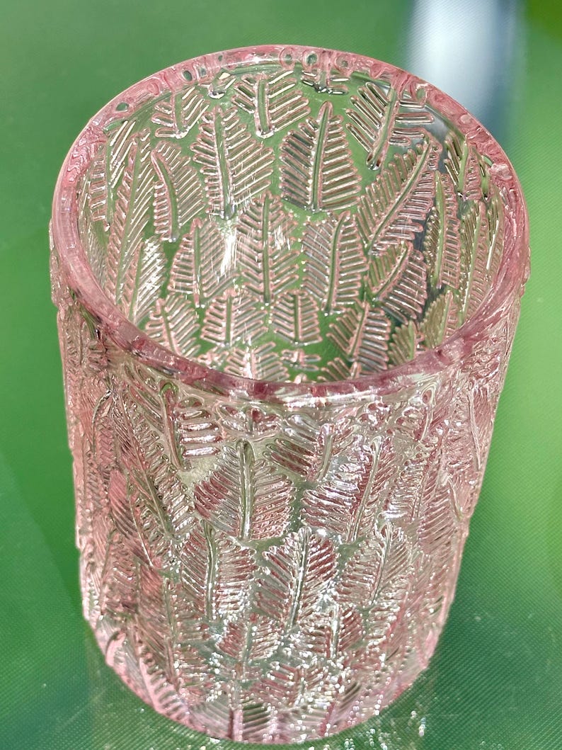 May include: A clear, pink glass tumbler with a detailed leaf pattern. The cylindrical glass has a slightly flared rim and is designed with a repeating embossed fern-like motif. The glass is suitable for beverages and decorative use.