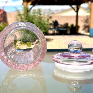 May include: A clear, pink glass jar with a matching lid. The jar is open, revealing its interior, and the lid is next to it. The lid has a round, clear knob on top. The jar and lid are on a reflective surface.