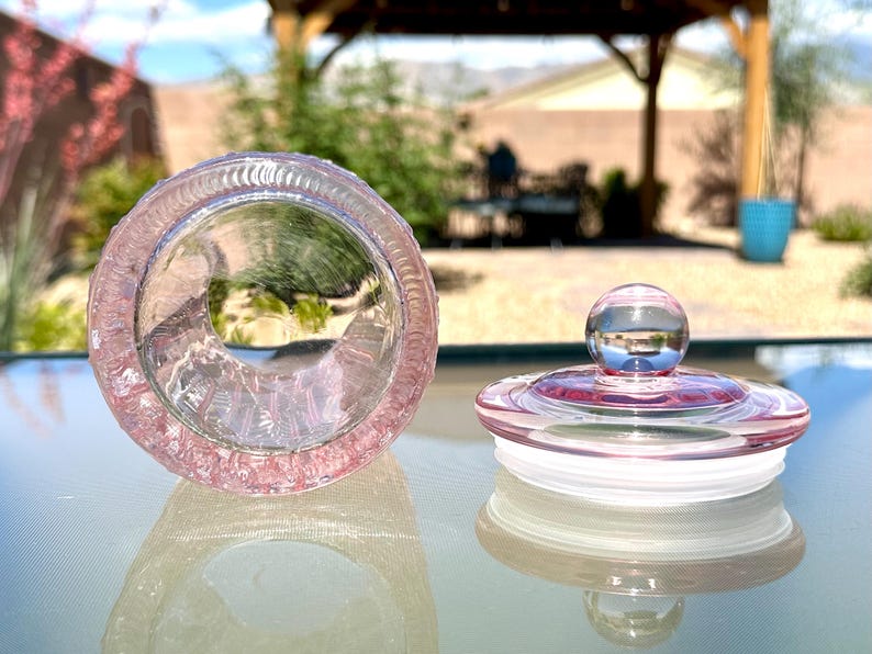 May include: A clear, pink glass jar with a matching lid. The jar is open, revealing its interior, and the lid is next to it. The lid has a round, clear knob on top. The jar and lid are on a reflective surface.
